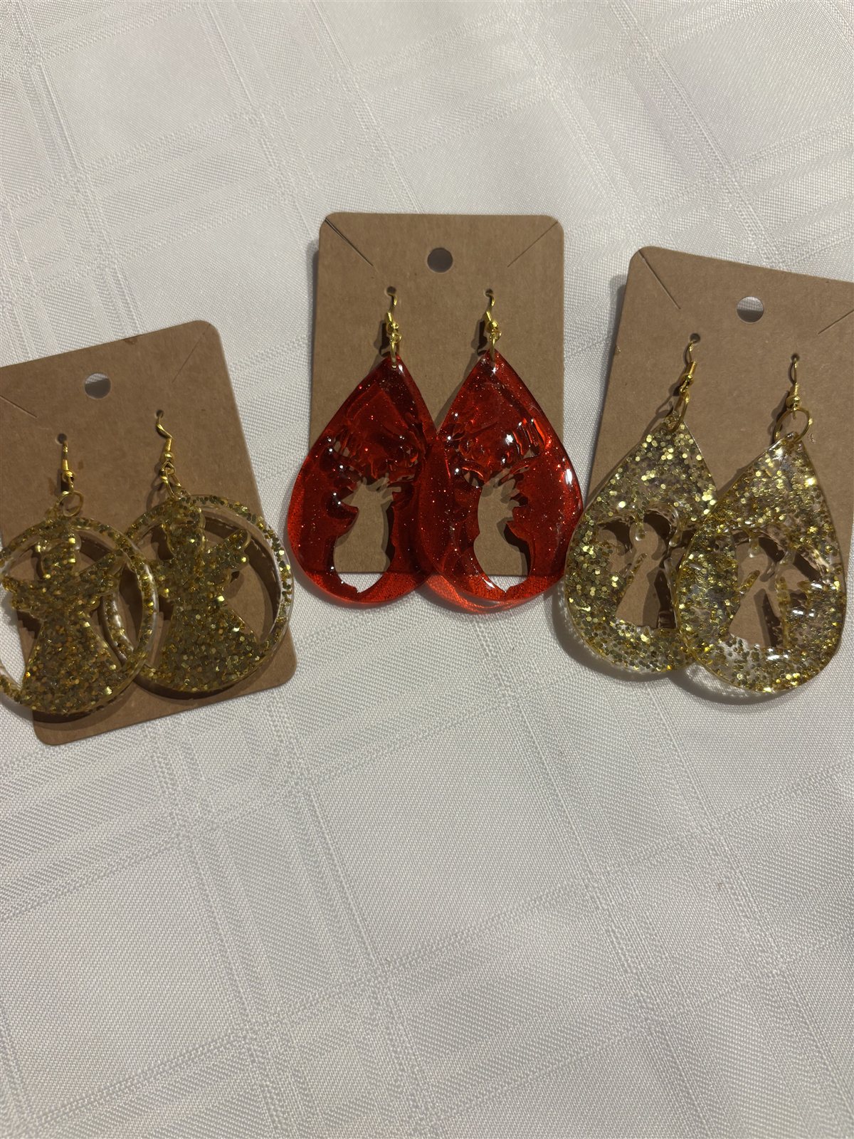 Three pairs of teardrop resin earrings on cards, each with an angel cut-out, in gold glitter, red and clear gold glitter