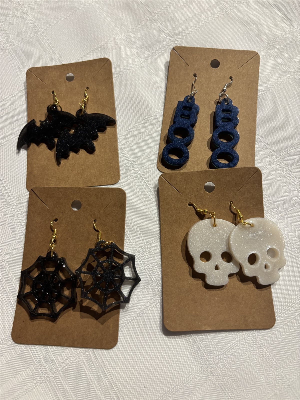 Four pairs of Halloween-themed resin earrings on cards, a bat, the word BOO, a spiderweb and a skull