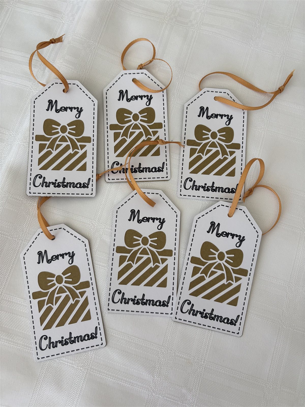Six white gift tags with a gold gift-box design and tan ribbon, reading Merry Christmas