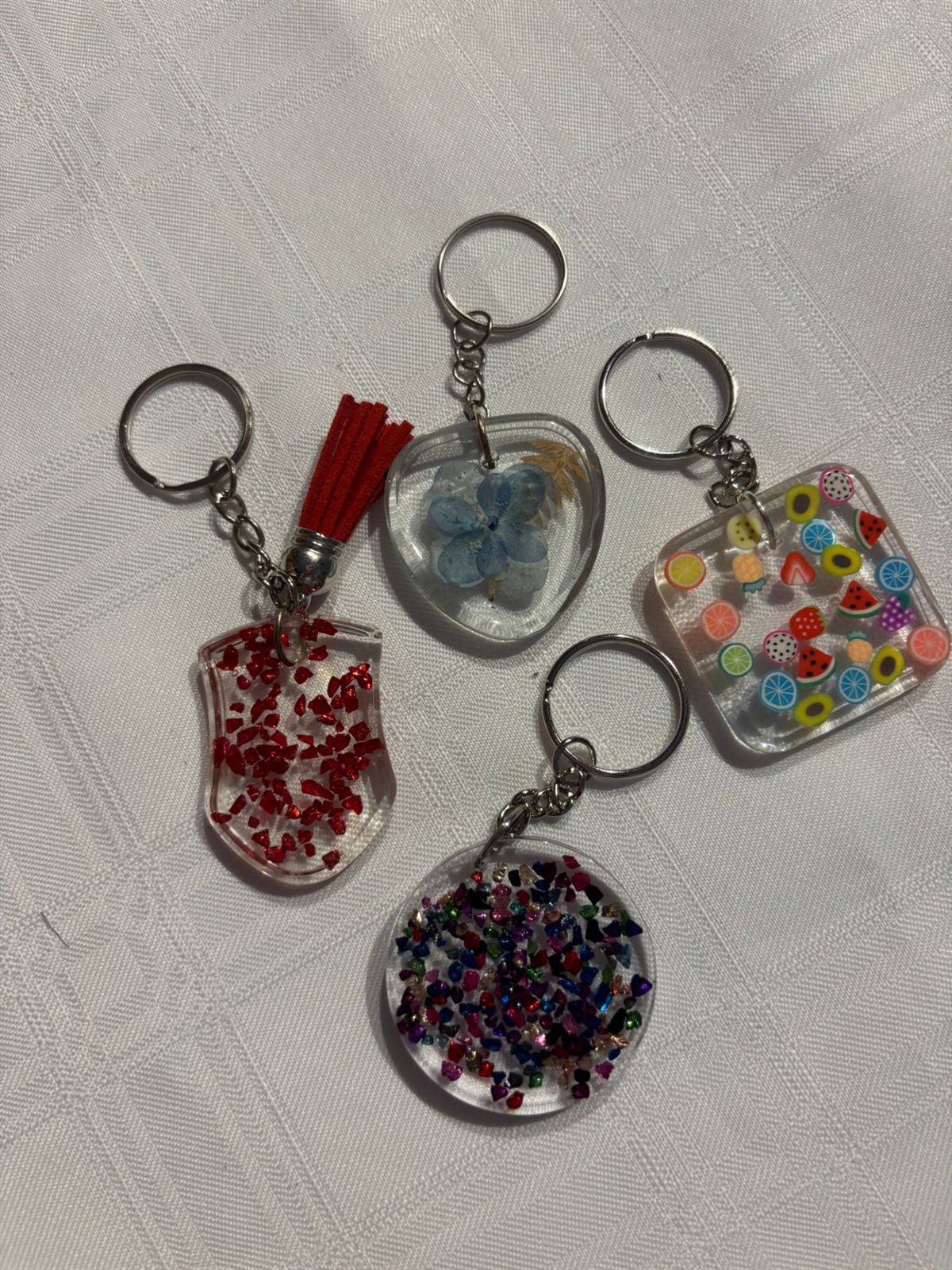 Four resin keychains, a red glitter shield with a tassel, a heart with a pressed blue flower, a square with fruit slices and a round one with coloured glitter